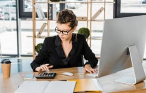 A focused professional in glasses using a calculator and computer at an office desk.