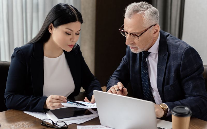 Two professionals in business attire engage with a laptop, discussing filing instructions related to export documentation.