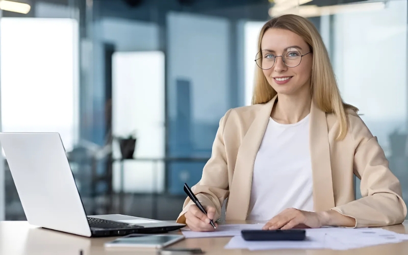 A woman with glasses is at her desk, engaged with her laptop, while reviewing IC-DISC compliance materials.