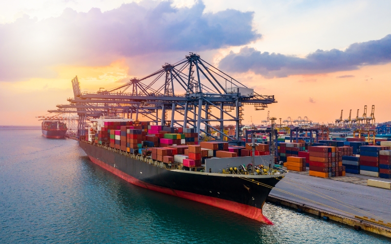 A large container ship sails on the water during a vibrant sunset, casting reflections on the calm sea surface.