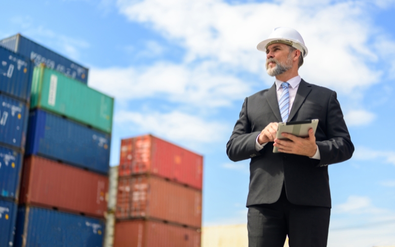 A suited man with a hardhat poses in front of shipping containers, representing the role of IC-DISC for U.S. exporters.