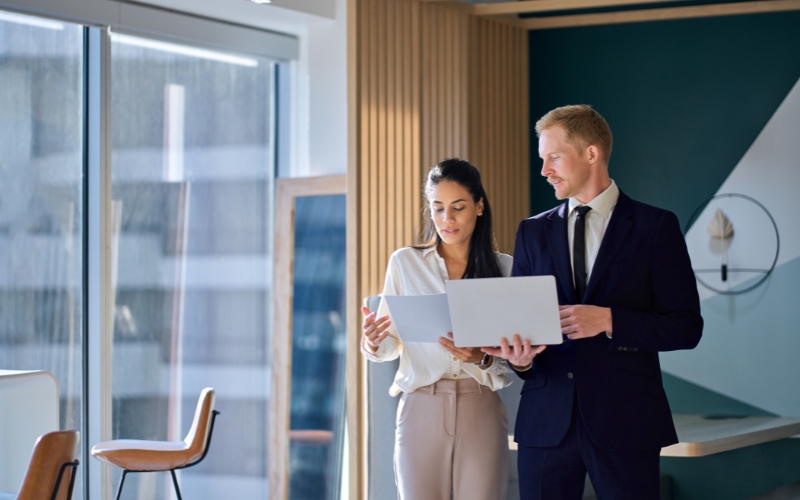 Two business individuals standing in an office, discussing the 1120-IC-DISC Schedule K document regarding compliance reminders.