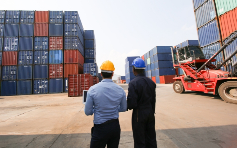 Two men are positioned before shipping containers, analyzing the advantages of IC-DISC for various industries and cost savings.