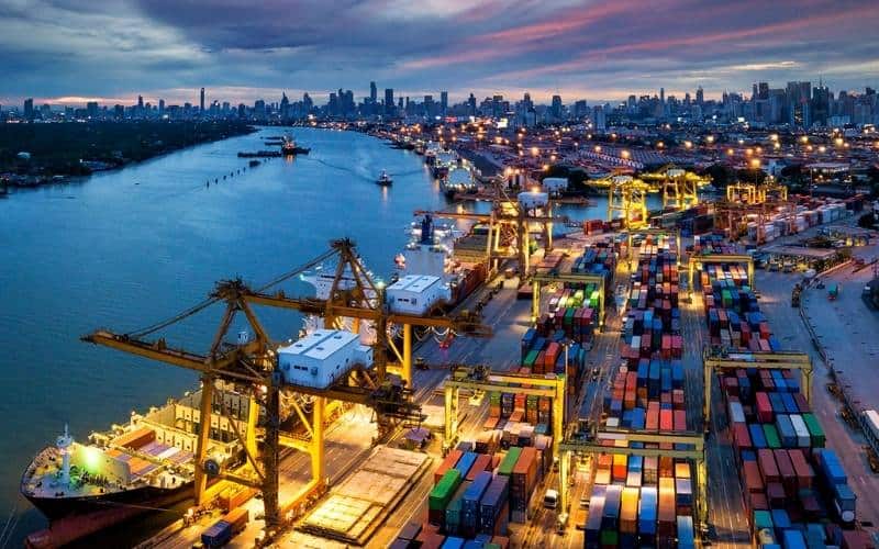 Aerial view of a port at dusk, showcasing ships and cranes against a colorful sky, highlighting the bustling maritime activity.