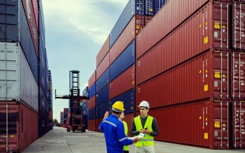 A group of professionals stand beside shipping containers, discussing the IC-DISC Commission Payment Rules and the 90-Day Rule.