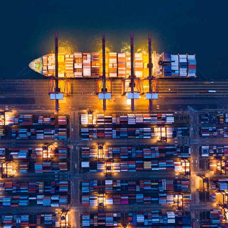 port Aerial view of a cargo ship being loaded with containers at a brightly lit port at night.