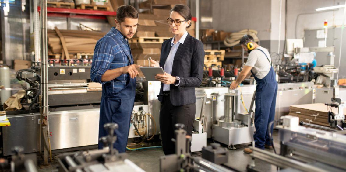 A manager and a factory worker discussing data on a tablet in a manufacturing facility.