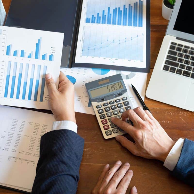 Overhead view of hands using a calculator and reviewing bar charts and data on a wooden desk.
