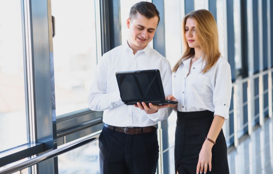 Two colleagues in professional attire discussing information on a laptop in a bright office hallway.